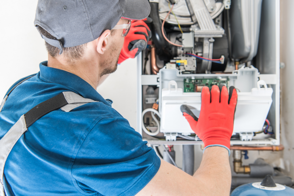 A technician wearing red gloves and a cap works on the open control panel of a wall-mounted heating system or boiler.
