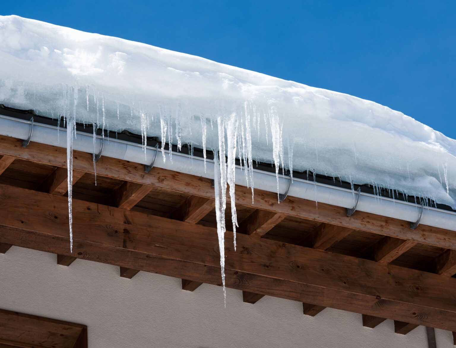 Icicles hang from the edge of a snow-covered roof under a clear blue sky, resembling delicate articles of winter’s artistry.