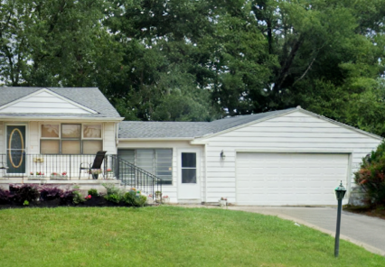 A single-story white house with a front porch, black railing, attached garage, and a green lawn bordered by trees in the background is featured in several articles highlighting charming home designs.