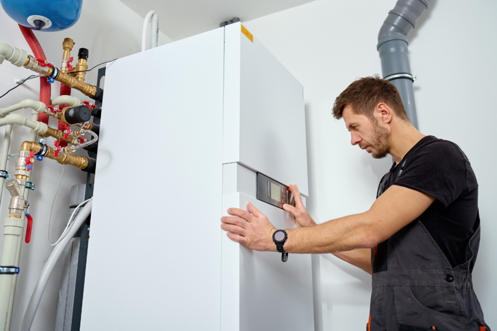A technician adjusts the control panel of a modern heating system unit in a utility room surrounded by pipes and valves.