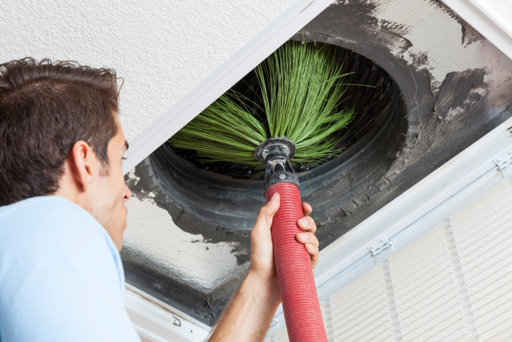 A worker uses a hose with a brush attachment to clean the inside of an air duct in a ceiling vent.