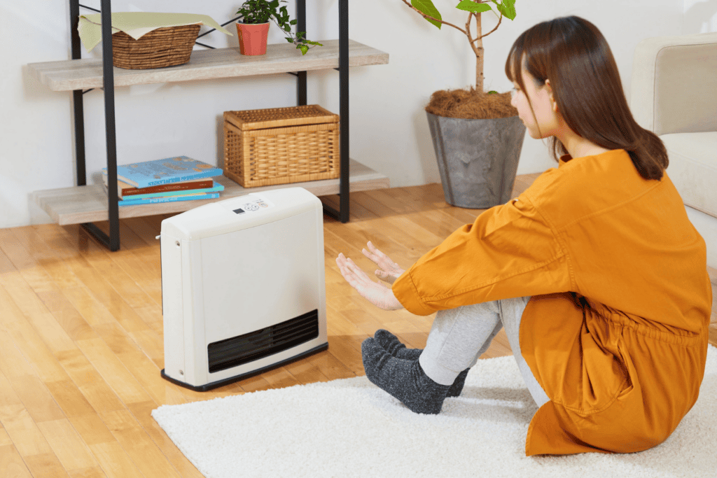 A woman in an orange dress sits on a rug, warming her hands and feet in front of a white electric heater in a living room.