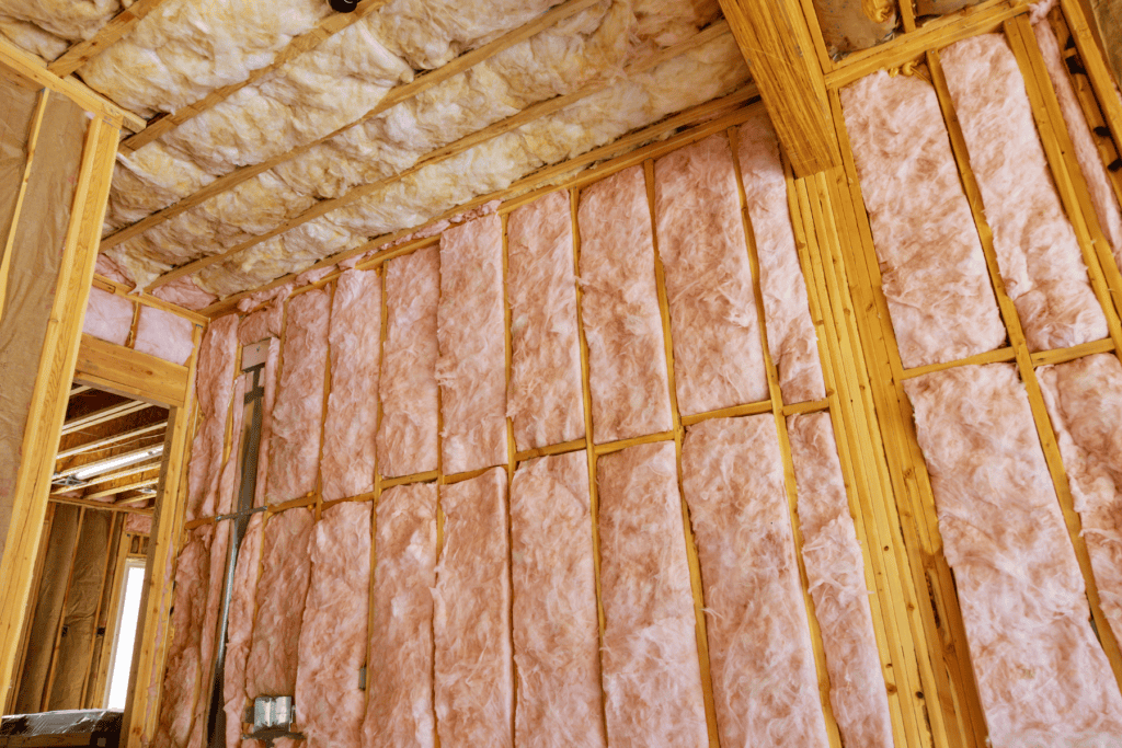 Pink fiberglass insulation is installed between wooden wall and ceiling studs in a building under construction.