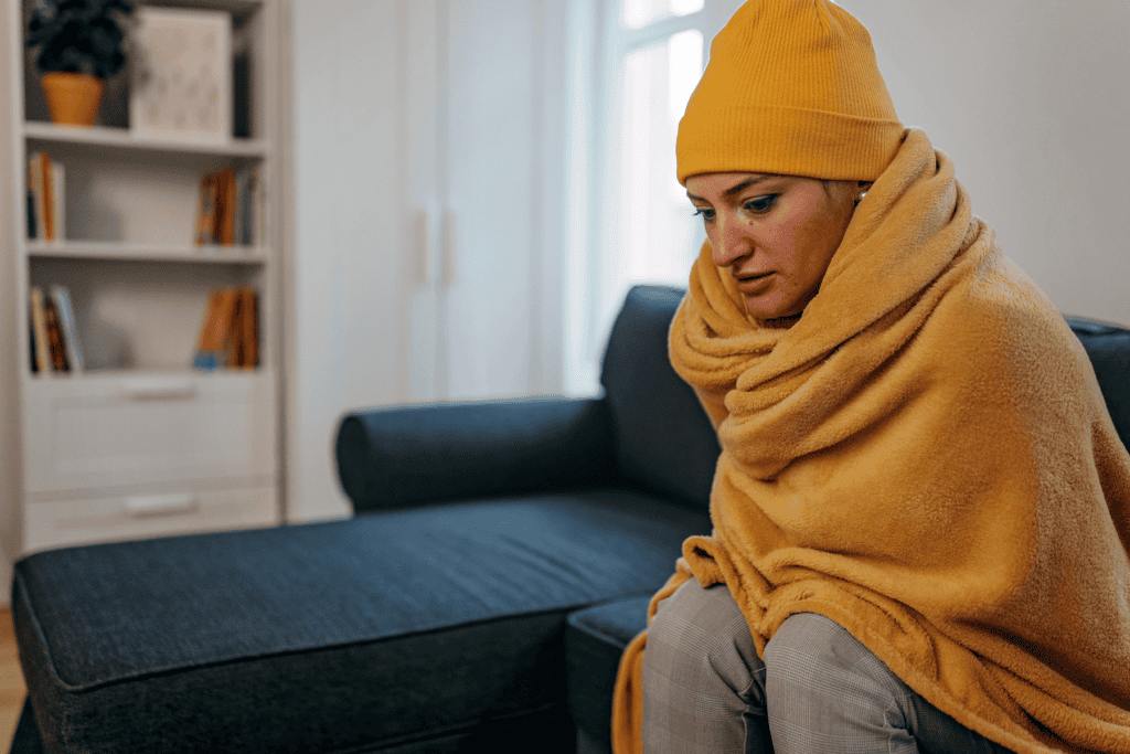 A person wearing a yellow hat and wrapped in a yellow blanket sits on a couch in a living room, appearing cold and thoughtful.