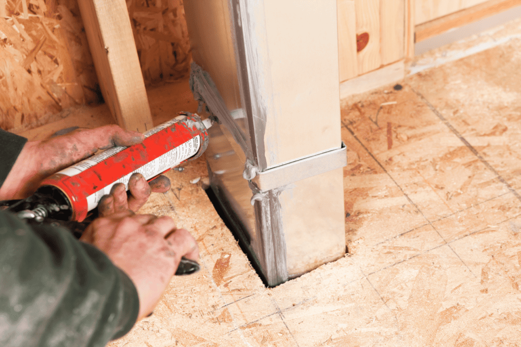 A person applies sealant around a metal duct where it passes through a plywood floor in a construction setting.